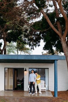 two people standing in front of a white building with blue roof and trees behind them