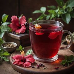 a glass cup filled with red liquid next to some flowers and herbs on a wooden table