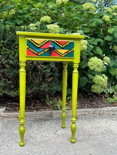 a green table with a colorful painted drawer on it's side in front of some bushes