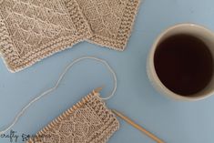 a cup of coffee next to two knitted coasters and a knitting needle on a blue surface