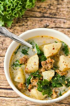 a bowl of soup with meat, potatoes and spinach on a wooden table next to a leafy green plant