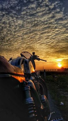 a motorcycle parked on the side of a dirt road at sunset with clouds in the sky