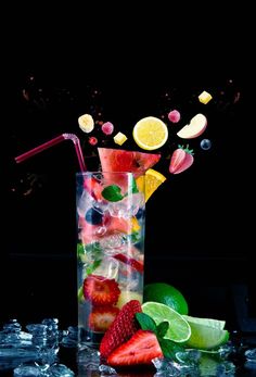 a tall glass filled with fruit and ice on top of a black table next to lemons, strawberries, limes, and watermelon