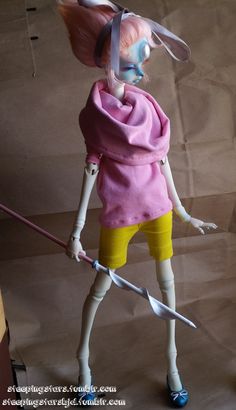 a doll with pink hair and yellow shorts holding a stick in one hand while standing on top of a brown paper bag