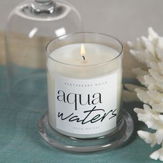 an aqua waters candle sitting next to a white flower on a blue table cloth with a clear glass vase behind it