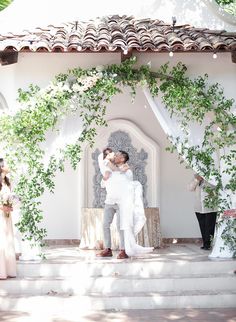 a bride and groom kissing in front of an outdoor wedding ceremony arch with greenery