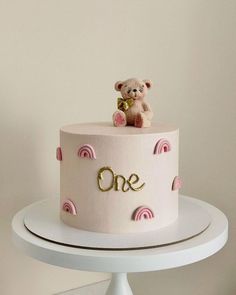 a one tiered cake with a teddy bear on top and the word one written in gold