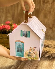 a hand holding a paper house with a rabbit on the front and windows painted on it