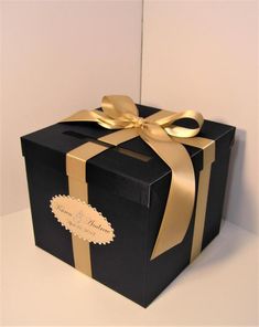 a black box with gold ribbon and a bow on it's side, sitting on a white surface