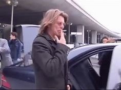 a man standing in front of a car talking on a cell phone while holding his hand to his ear