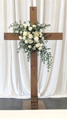 a wooden cross with white flowers and greenery on it in front of a curtain