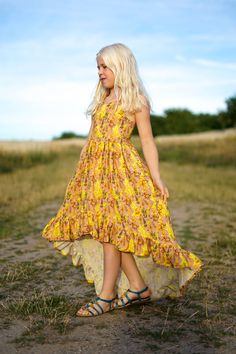 a woman in a yellow dress is standing on a dirt road and looking off into the distance