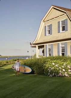 a man holding a surfboard in front of a house with flowers and bushes around it