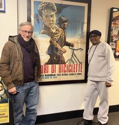 two men standing next to each other in front of a wall with posters on it