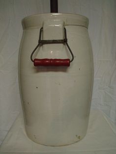 an old white jar with a red handle on the bottom and a metal hook in the middle