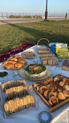 a table full of sandwiches and breads on a picnic blanket in the grass near a park