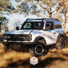 a white ford bronco is parked in the middle of some dry grass and trees