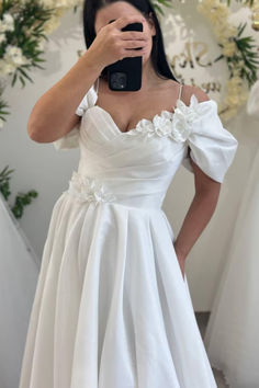 a woman taking a selfie wearing a white wedding dress with flowers on the shoulder