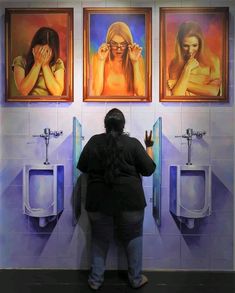 a woman standing in front of two urinals with paintings on the wall behind her