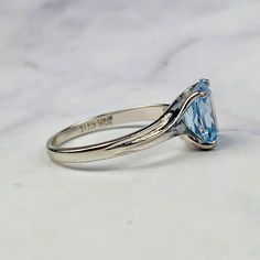 Beautiful vintage Aquamarine solitaire ring in 10K white gold. Emerald cut Aquamarine stone is set in a four prong setting on asymmetric shoulder resembling a bypass ring. Further details are as follows: Aquamarine - approximately 8mm x 6.4mm Total weight: 1.812 grams Metal: 10K white gold Stone: Emerald cut aquamarine Ring Size: 5.5 US, K/L UK Marking: 10K, GTR Makers Mark (Samuel Aaron) Condition: Excellent Ring is currently size 5.5 US and resizable. Please inquire for a quote. Thank you for Classic Blue Topaz Birthstone Ring, Classic Blue Topaz Birthstone Ring, Round Cut, Classic Round Cut Blue Topaz Birthstone Ring, Classic Birthstone Ring With Round Cut Blue Topaz, Classic Emerald Cut Topaz Ring With Prong Setting, Classic Round Bypass Ring With Tension Setting, Classic Bypass Ring With Diamond Cut, Classic Bypass Ring With Round Diamond Cut, Classic Bypass Ring With Solitaire