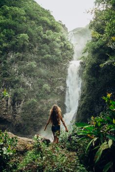 a woman standing in front of a waterfall with her back to the camera, looking down