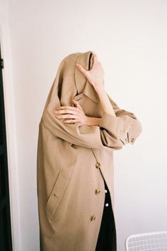 a woman in a trench coat is covering her face with one hand while standing against a wall