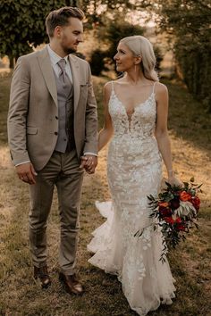 a bride and groom holding hands walking through the grass in front of some trees at sunset