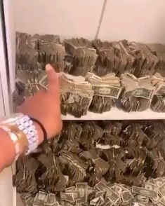 a person is pointing at stacks of money in a storage area with white walls and shelves