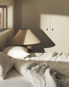 an unmade bed with white linens and a lampshade in the corner