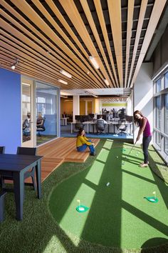 two women playing mini golf in an open office space with wood slats on the ceiling