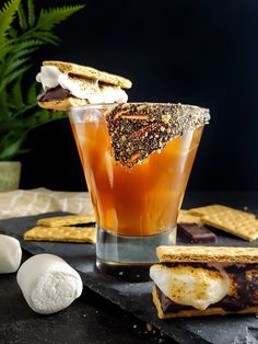 a drink and some crackers on a black table with plants in the background,