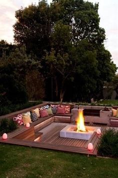 an outdoor fire pit surrounded by wooden decking and pillows on the ground with trees in the background