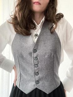 a woman wearing a gray vest and white shirt with black pleated skirt standing in front of a wall