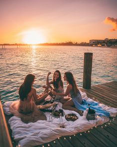 three women sitting on a dock talking and having a picnic with the sun setting in the background