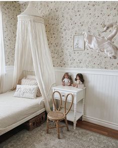 two dolls sitting on a small white bed in a room with floral wallpaper and curtains