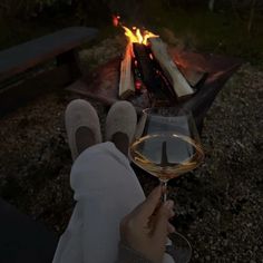 a person holding a glass of wine in front of a campfire with their feet up