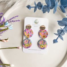 a pair of colorful earrings sitting on top of a white plate next to blue flowers