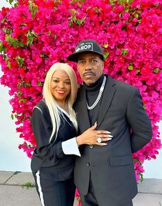 a man and woman standing next to each other in front of pink flowers with their arms around each other