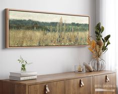 a painting hanging on the wall above a dresser with flowers and books in front of it