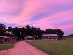 a dirt road in front of some wooden buildings and trees at dusk with pink clouds