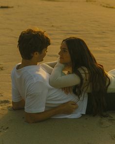 a man and woman sitting on the beach together looking at each other's eyes