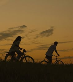 two people riding bikes in the grass at sunset