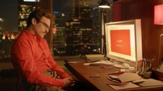 a man sitting at a desk in front of a computer with the city lights behind him