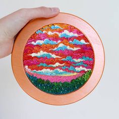 a hand is holding up a colorful painting on an orange plate with clouds in the sky