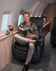 a man is sitting in an airplane reading a book and smiling at the camera,