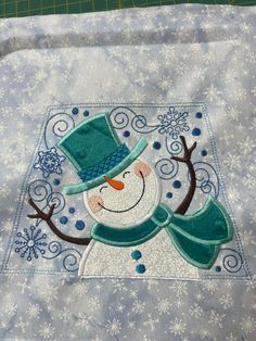 a snowman with a green hat and scarf on top of a quilted blanket