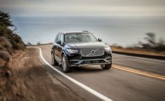 the front end of a black volvo suv driving down a road near the ocean with mountains in the background