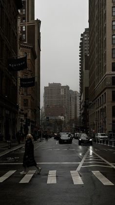 a woman walking across a street in the middle of a crosswalk with cars and buildings behind her