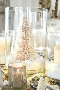 a table topped with glass vases filled with snow covered candles and a small christmas tree