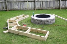 an outdoor fire pit surrounded by wooden raised planters and tools in the grass near a fence